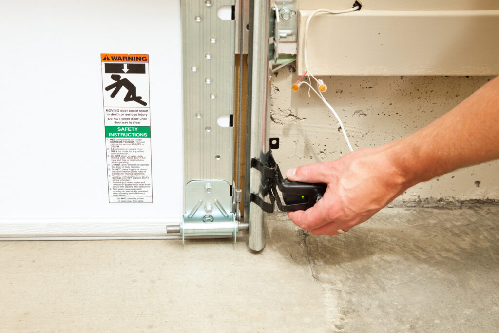 "A hand is adjusting a garage door safety reversing sensor, with an illuminated green LED to confirm the sensor is lined up with the opposite side sensor and operational. These sensors, required in all new installations, reverse the door when an infrared beam is broken by a child, animal or object. A warning and safety instructions sticker is to the left."