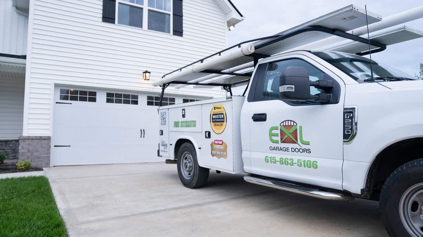 A truck labeled "EXL Garage Doors" is parked in front of a white house with a double garage door. The truck carries supplies on its roof rack.