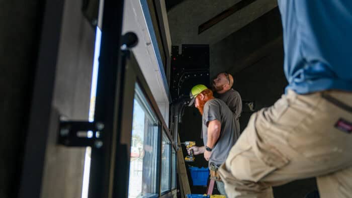 Three workers wearing casual workwear and safety gear are installing or adjusting equipment near a large window inside a building.